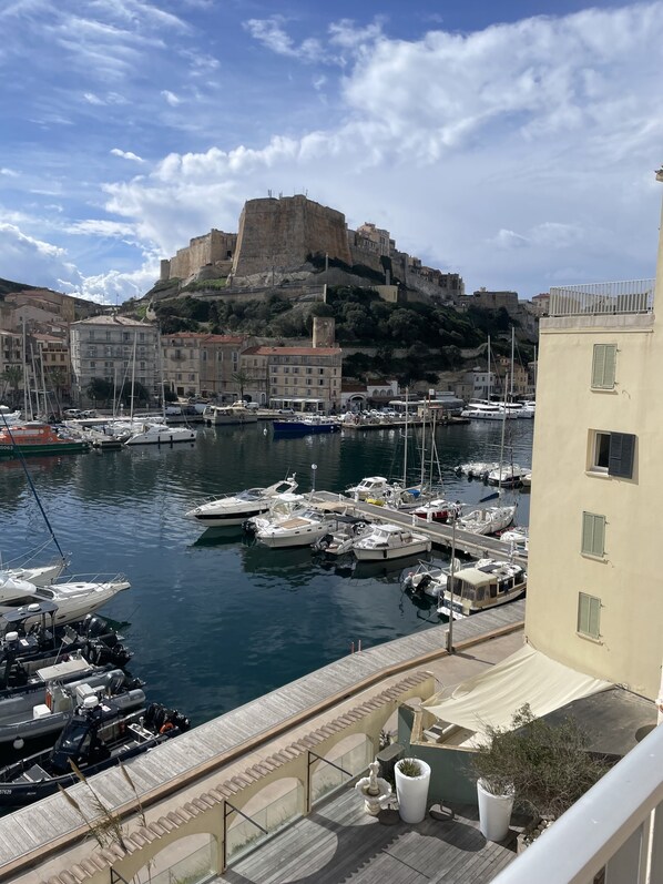 A marina with boats docked, a historic fort on a hill, and buildings with balconies.