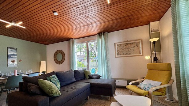 Un salon avec un plafond en bois, un canapé, un fauteuil jaune, une petite table et une fenêtre avec des rideaux.