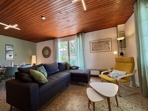 Un soggiorno con soffitto in legno, un divano, una poltrona gialla, un tavolino e una finestra con tende.