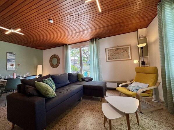 A living room with a wooden ceiling, a sofa, a yellow armchair, a small table, and a window with curtains.
