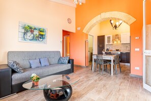 A living room with a sofa, a glass coffee table, a dining area with chairs, and a kitchen visible through an archway.