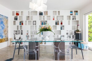 A modern dining area with a glass table, clear chairs, and a white bookshelf filled with books and decorative items. - Villa Marianna - Terrasini (Terrasini)