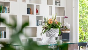 A modern interior with a glass table, white shelving with books and decorative items, and a potted plant with flowers. - Villa Marianna - Terrasini (Terrasini)