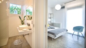 A modern bathroom with a white sink, mirror, and potted plants. It leads to a bedroom with a bed, bedside table, and a chair. - Villa Marianna - Terrasini (Terrasini)