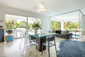 Une cuisine moderne dotĂ©e dâun Ăźlot central en verre, de chaises transparentes, dâun canapĂ© et offrant une vue sur une piscine et un jardin.