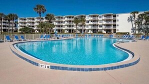 Una piscina con una profundidad de 99 centímetros, rodeada de sillas reclinables azules y palmeras, con hoteles de varios pisos al fondo.