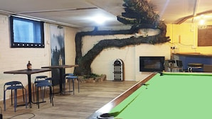 A pool table in a room with a brick wall, a bar area with shelves of bottles, and a television mounted on the wall.