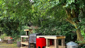 A portable outdoor cooking station with a grill, a red cooler, and a trash bin, set in a wooded area.