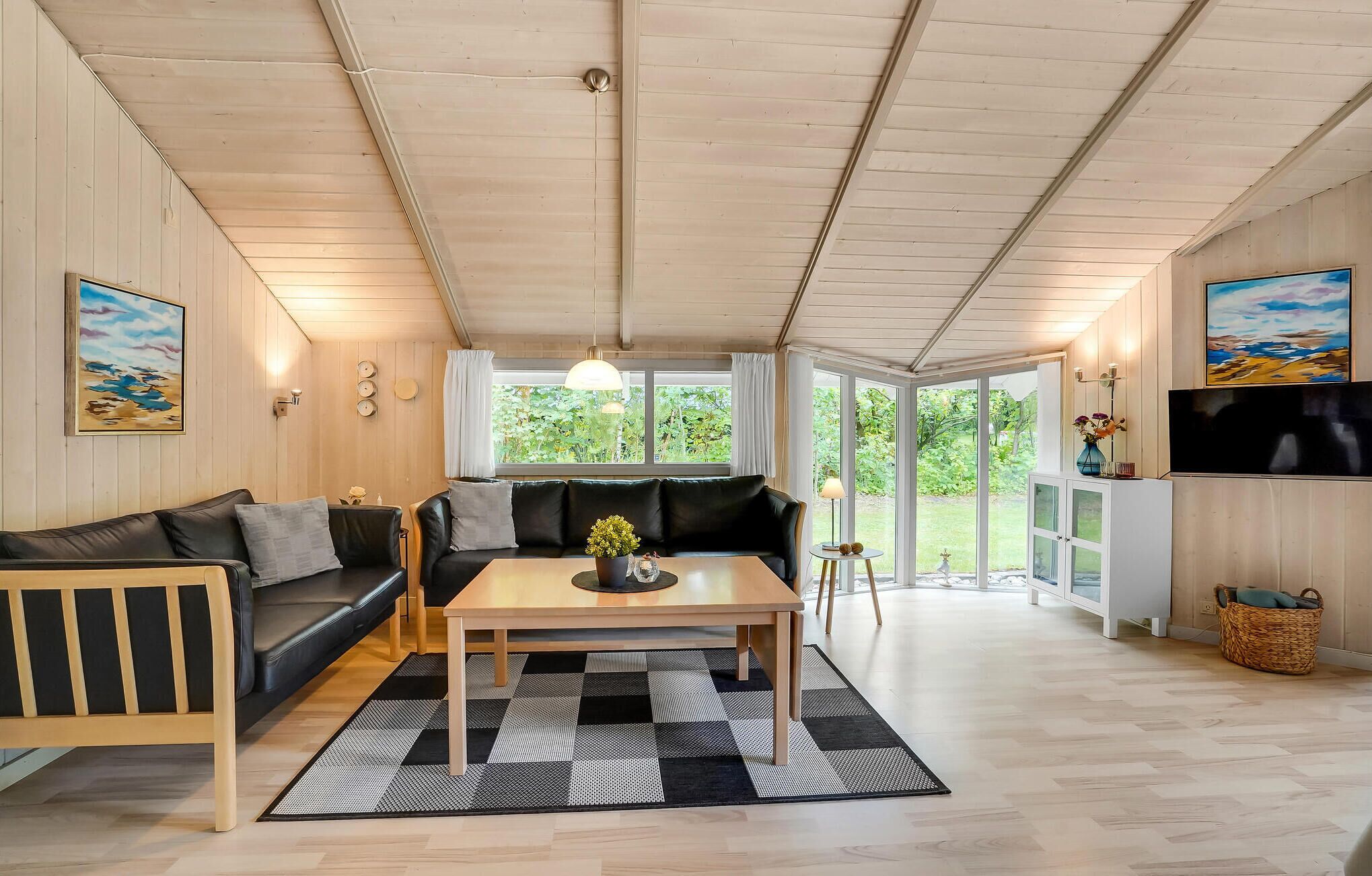 Ein gemütliches Wohnzimmer mit einer schwarzen Ledersofa, einem Holztisch und einem karierten Teppich. Große Fenster bieten Blick auf Grünflächen außerhalb.