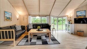 Un salón acogedor con un sofá de cuero negro, una mesa de centro de madera y una alfombra a cuadros. Amplias ventanas ofrecen vistas al verdor exterior.