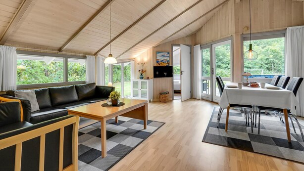 Amplia sala de estar con sofá, mesa de centro y comedor, ventanas grandes con vistas a zonas verdes, y una cocina con televisor.