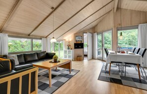 Ett rymligt vardagsrum med en soffa, ett soffbord och ett matbord, stora fönster med utsikt över grönområden samt ett köksområde med en tv.
