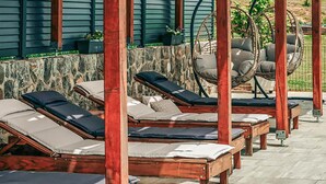 Une rangée de chaises longues en bois, dotées de coussins, est placée devant un mur de pierre et un bâtiment vert aux bardages horizontaux.