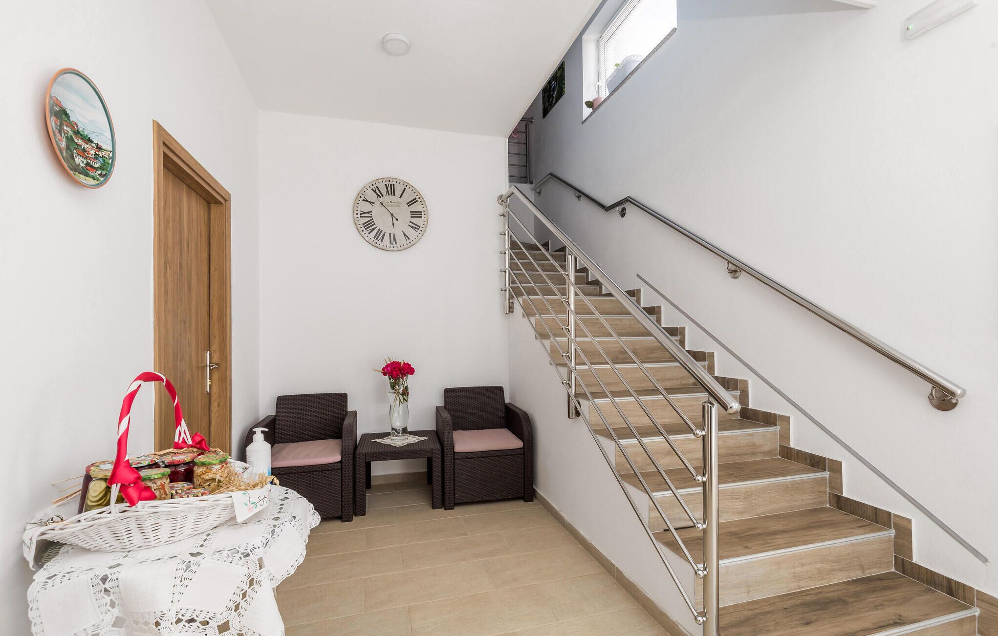 Eine Treppe mit Metallgeländer, eine Holztür, zwei Korbstühle, ein kleiner Tisch mit weißer Tischdecke und eine Wanduhr.