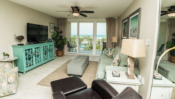 Een woonkamer met een televisie, een blauwgroene kast, bank, poef en uitzicht op de oceaan door grote ramen.