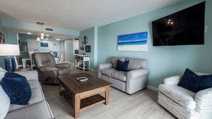 A living room with a sofa, a coffee table, a TV, and a painting of a beach scene. - D406 Corner Unit, Great View of Pool and Direct Gulf View (Panama City Beach)