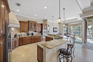A spacious kitchen with wooden cabinets, a large island, and a dining area with bar stools. - Opulent Waterfront Home | Pool, Spa, Dock & Deck (Marco Island)