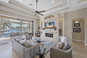 A spacious living room with a large TV, comfortable seating, a fireplace, and a view of a pool. - Opulent Waterfront Home | Pool, Spa, Dock & Deck (Marco Island)