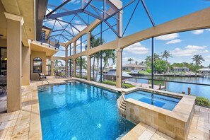 A swimming pool with a glass bottom, surrounded by a patio area with lounge chairs and a view of a body of water and palm trees. - Opulent Waterfront Home | Pool, Spa, Dock & Deck (Marco Island)