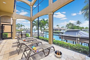 A covered outdoor patio with lounge chairs, a grill, and a view of a marina with boats. - Opulent Waterfront Home | Pool, Spa, Dock & Deck (Marco Island)