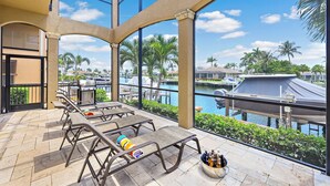 A covered outdoor patio with lounge chairs, a grill, and a view of a marina with boats.