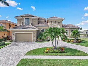 A two-story house with a garage, a covered patio, and a landscaped front yard with palm trees and flowers. - Opulent Waterfront Home | Pool, Spa, Dock & Deck (Marco Island)