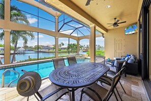 An outdoor pool area with a table and chairs, a ceiling fan, and a view of a body of water and palm trees. - Opulent Waterfront Home | Pool, Spa, Dock & Deck (Marco Island)