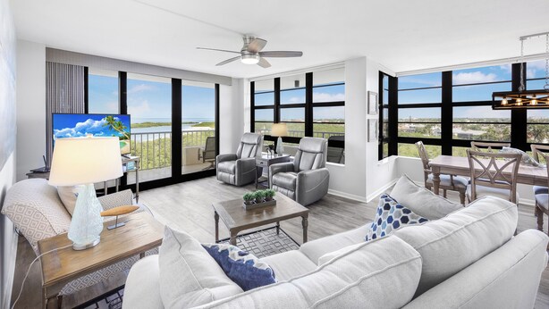 A spacious living area with a sofa, coffee table, and chairs, large windows with a view, and a ceiling fan. - Corner Beachfront Condo | Gym, Pool & Grills (Marco Island)