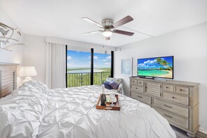 A bedroom with a large bed, a ceiling fan, a television, a dresser, and a balcony with a view.