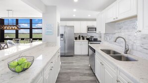 A modern kitchen with white cabinets, stainless steel appliances, and a large window overlooking a scenic view. - Corner Beachfront Condo | Gym, Pool & Grills (Marco Island)
