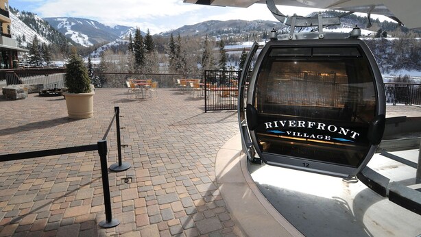 Un telesilla con letrero que dice 'Riverfront Village', un área pavimentada con bancos y una vista de montañas nevadas.