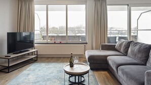 A modern living room with a flat-screen TV, a grey sofa, a round coffee table with a bottle and glasses, and large windows with curtains. - Spacious and luxurious apartment - Kaag Resort (27) (Warmond)
