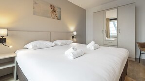 A neatly made bed with white linens and rolled towels, a bedside table with a lamp, a large wardrobe, and a painting on the wall. - Spacious and luxurious apartment - Kaag Resort (27) (Warmond)