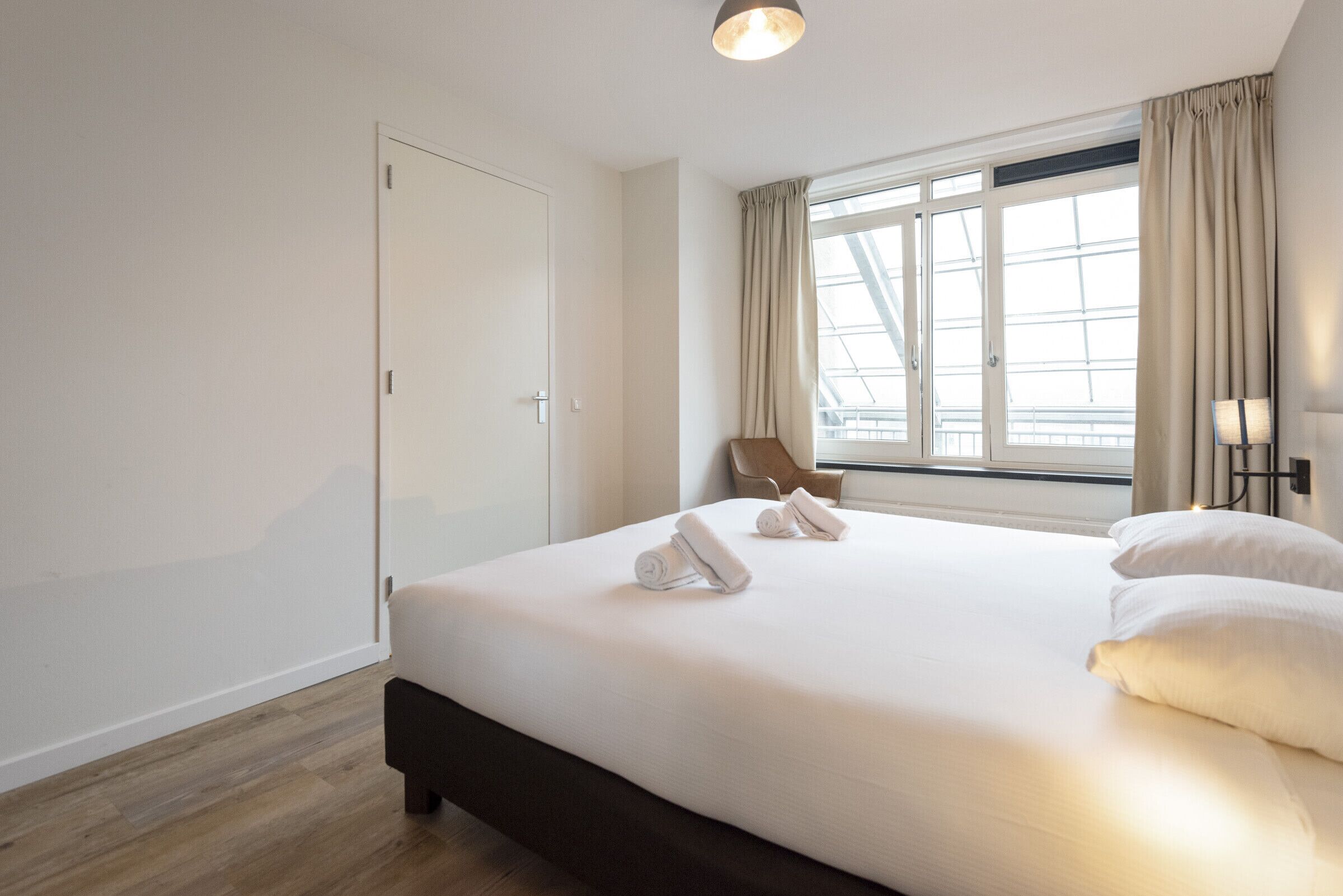 A hotel room with a large bed, a window with curtains, a chair, and a lamp.