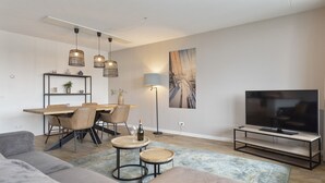 A modern living room with a sofa, a dining table with chairs, a TV stand, pendant lights, and a painting on the wall. - Spacious and luxurious apartment - Kaag Resort (27) (Warmond)