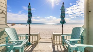 Strandudsigt fra en overdækket veranda med to Adirondack-stole og to stribede parasoller.