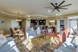 A spacious living room with a large coffee table, a dining area with wicker chairs, a kitchen with dark cabinets, and a ceiling fan.