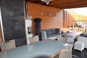 Een terras buiten met rieten meubels, een glazen tafel en een houten wand.