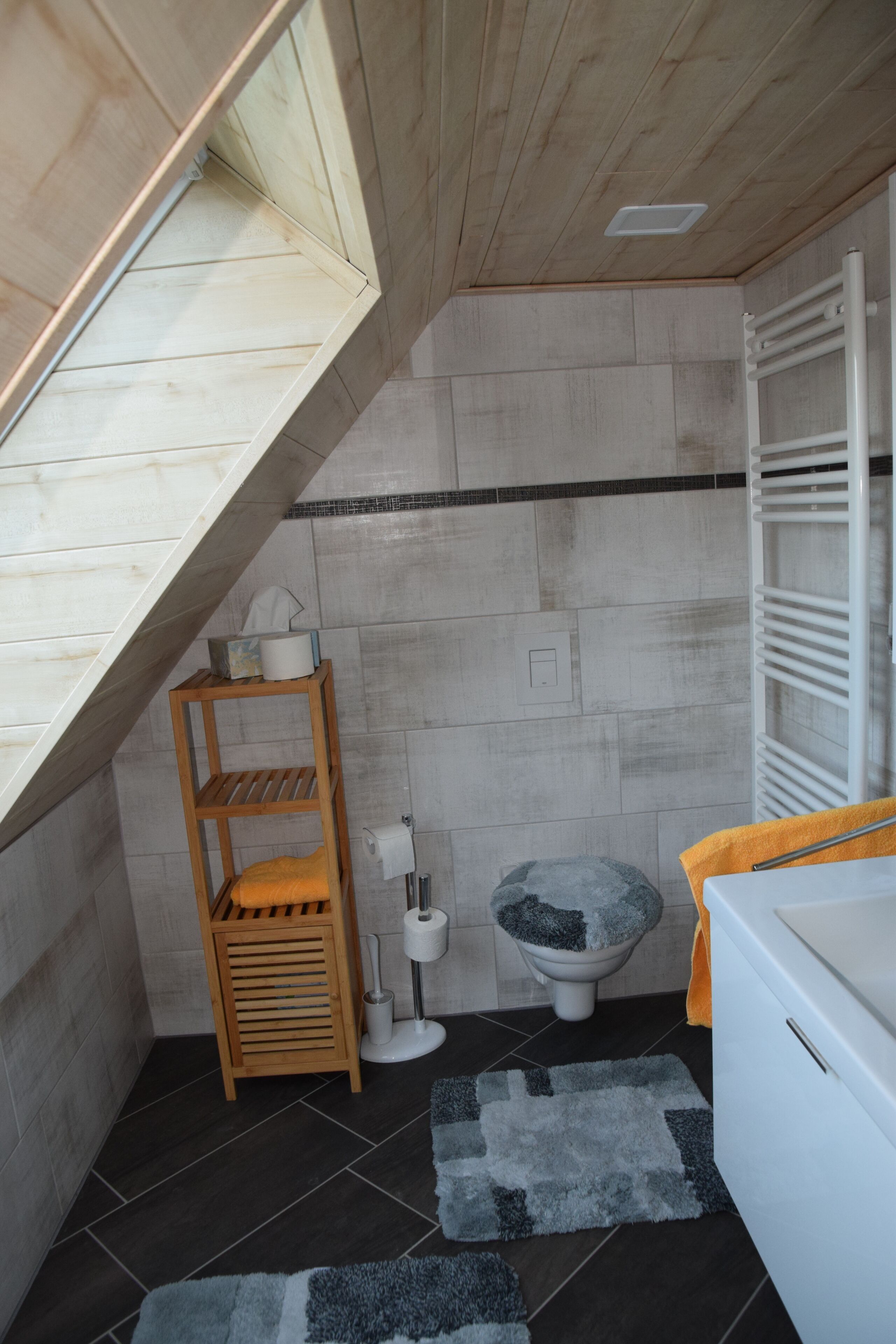 A compact bathroom with a sloped ceiling, a toilet, a small wooden shelf, and a white bathtub.