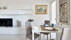 A dining area with a white table set for two, a television on a white cabinet, and a painting on the wall. - Sea view five minutes from the beach IUN Q5161 (Quartu Sant'Elena)