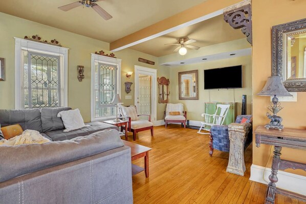 A living room with a grey sofa, wooden chairs, a television, and a ceiling fan.