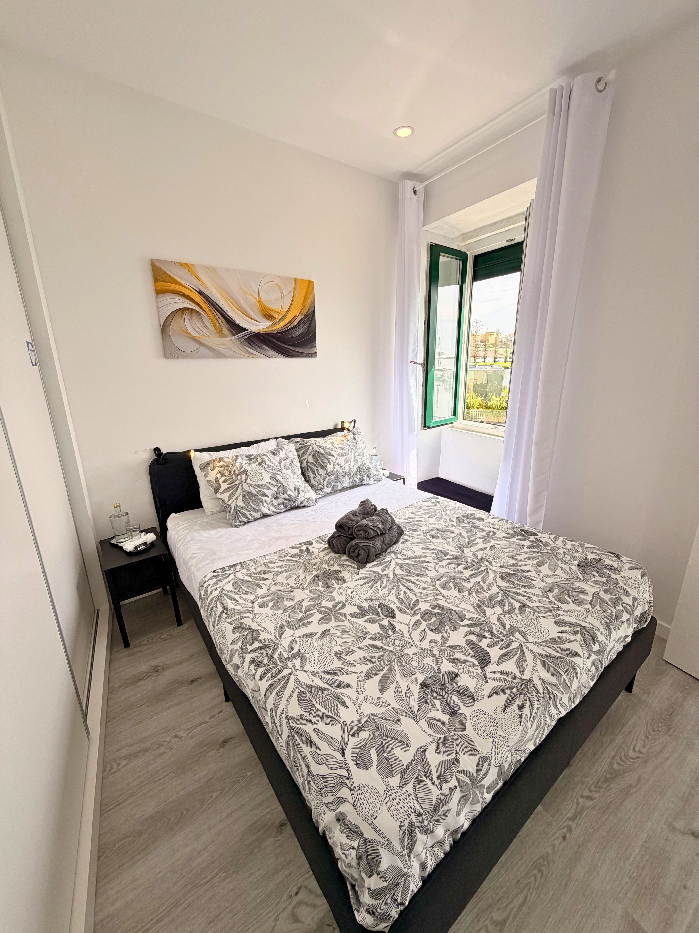 A bedroom with a bed, a nightstand, a window with curtains, and a wall-mounted abstract painting.