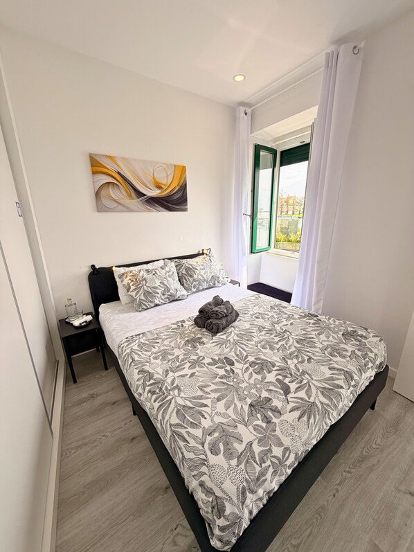 A bedroom with a bed, a nightstand, a window with curtains, and a wall-mounted abstract painting.