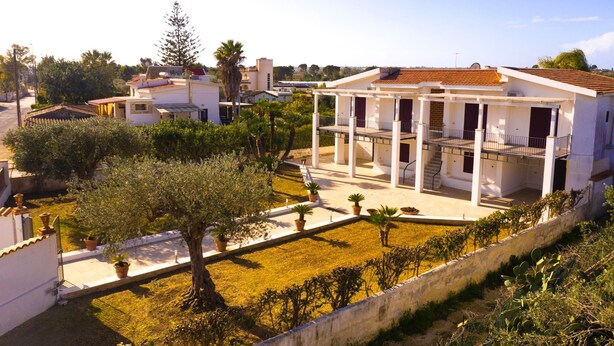 Una villa bianca con un portico coperto e un giardino con alberi di ulivo.