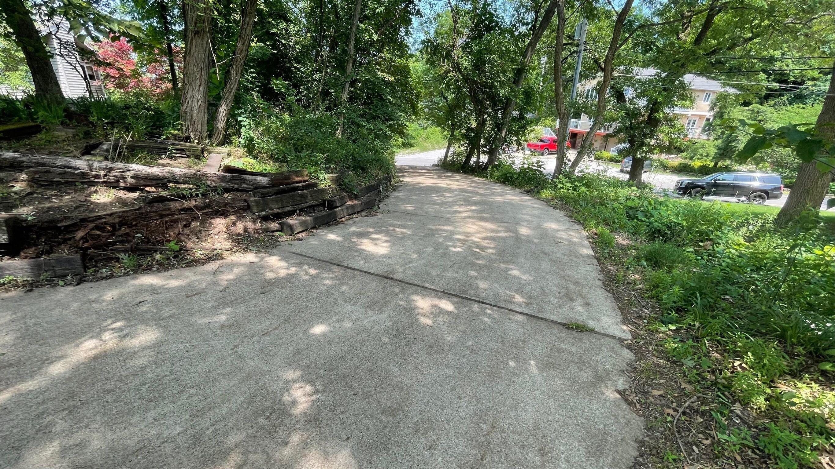 Ein Betonweg, gesäumt von Bäumen und Grünpflanzen, führt zu einem Wohngebiet mit parkenden Autos.
