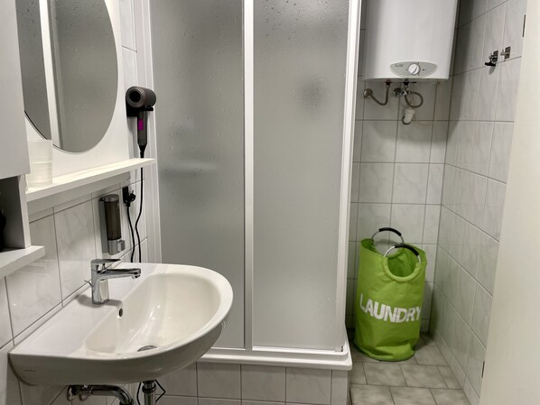 A bathroom with a white sink, a mirror, a shower enclosure, and a green laundry bag.