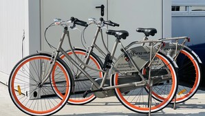 Ein Tretroller mit orangefarbenen und weißen Reifen steht vor einem Gebäude, an dem ein Schild mit der Aufschrift "Tuning-BUS" angebracht ist.