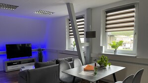 Ein modernes Wohnzimmer mit einem grauen Sofa, einem Fernseher, einem Esstisch mit Stühlen und einem Fenster mit Jalousien.