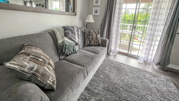 Un salon avec un canapé gris, un plaid à motifs et un tapis gris. Il y a un grand miroir au mur, une lampe et une fenêtre avec des rideaux.