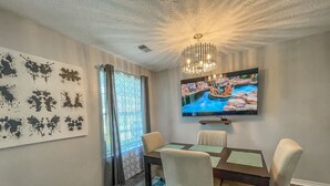 Un coin repas comprenant une table, des chaises, un lustre, un téléviseur et une fenêtre avec des rideaux.